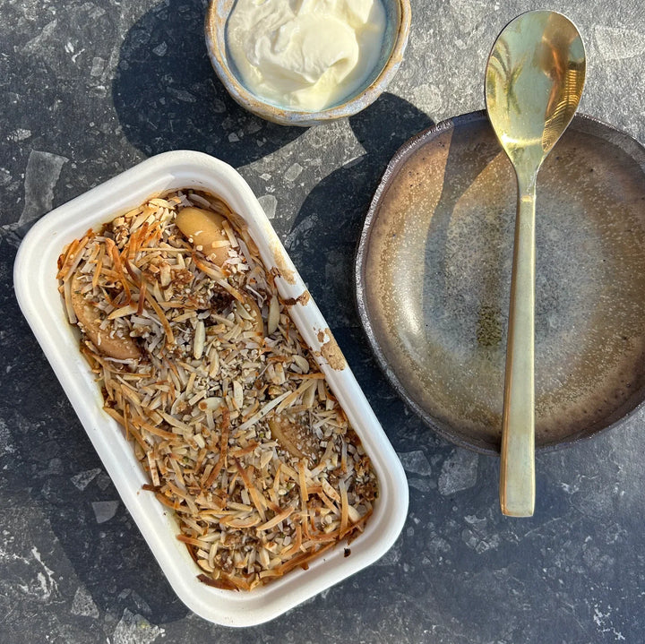 A container of apple crumble next to a ceramic plate with a spoon and a small rustic bowl with yoghurt