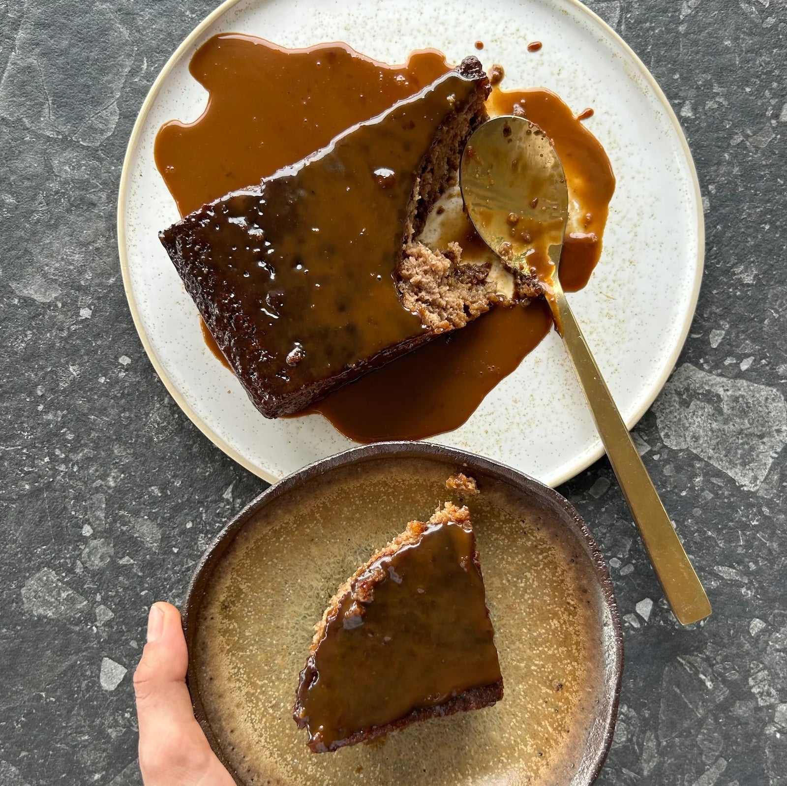 Hand holding slice of sticky date pudding with caramel sauce beside serving plate