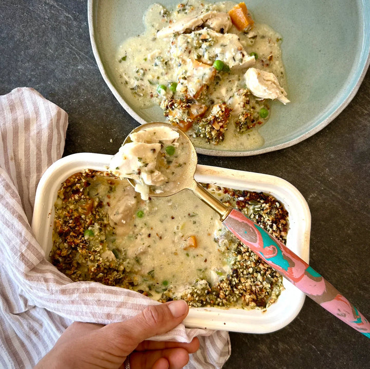A hand holding a spoonful of organic chicken and leek pie, with a plate of the same pie in the background.