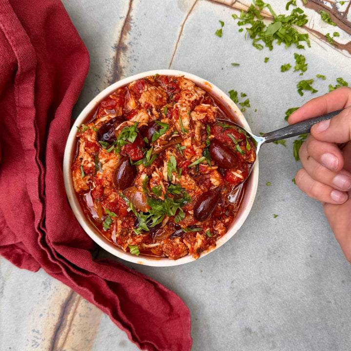 Bowl of chicken and olive stew with a spoon, garnished with herbs, on a textured surface with a red cloth.