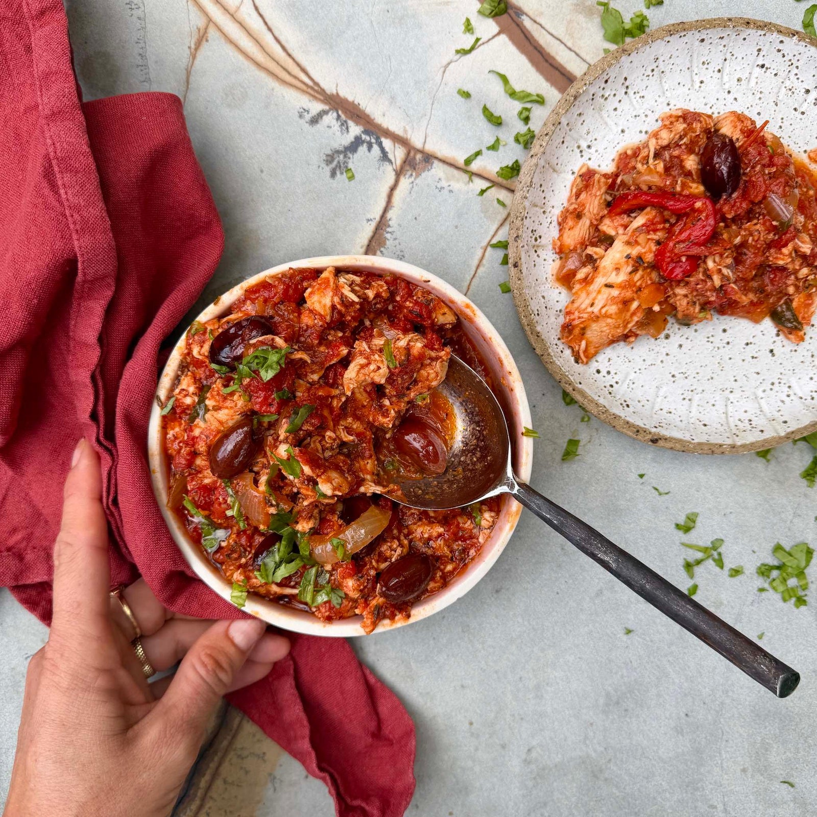 Bowl of hearty chicken and tomato stew with olives with a spoon, placed on a textured surface with a red cloth.